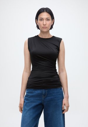 Donna con capelli corti e scuri che indossa un top nero arricciato senza maniche e jeans blu, in piedi davanti a uno sfondo bianco semplice.