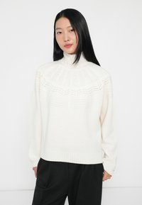 Pulôver branco de malha com gola alta, apresentando padrões texturizados e uma bainha canelada. Combinado com calças largas pretas.