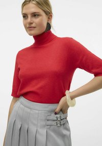 Maglione rosso a collo alto con maniche corte abbinato a una gonna plissettata grigio chiaro, con dettagli in metallo e un braccialetto traslucido.