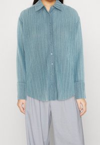 Niebieska koszula zapinana na guziki, plisowana, wykonana z miękkiego, teksturowanego materiału, z kołnierzykiem i długimi rękawami. Posiada pasujące guziki oraz zaokrąglony dół.