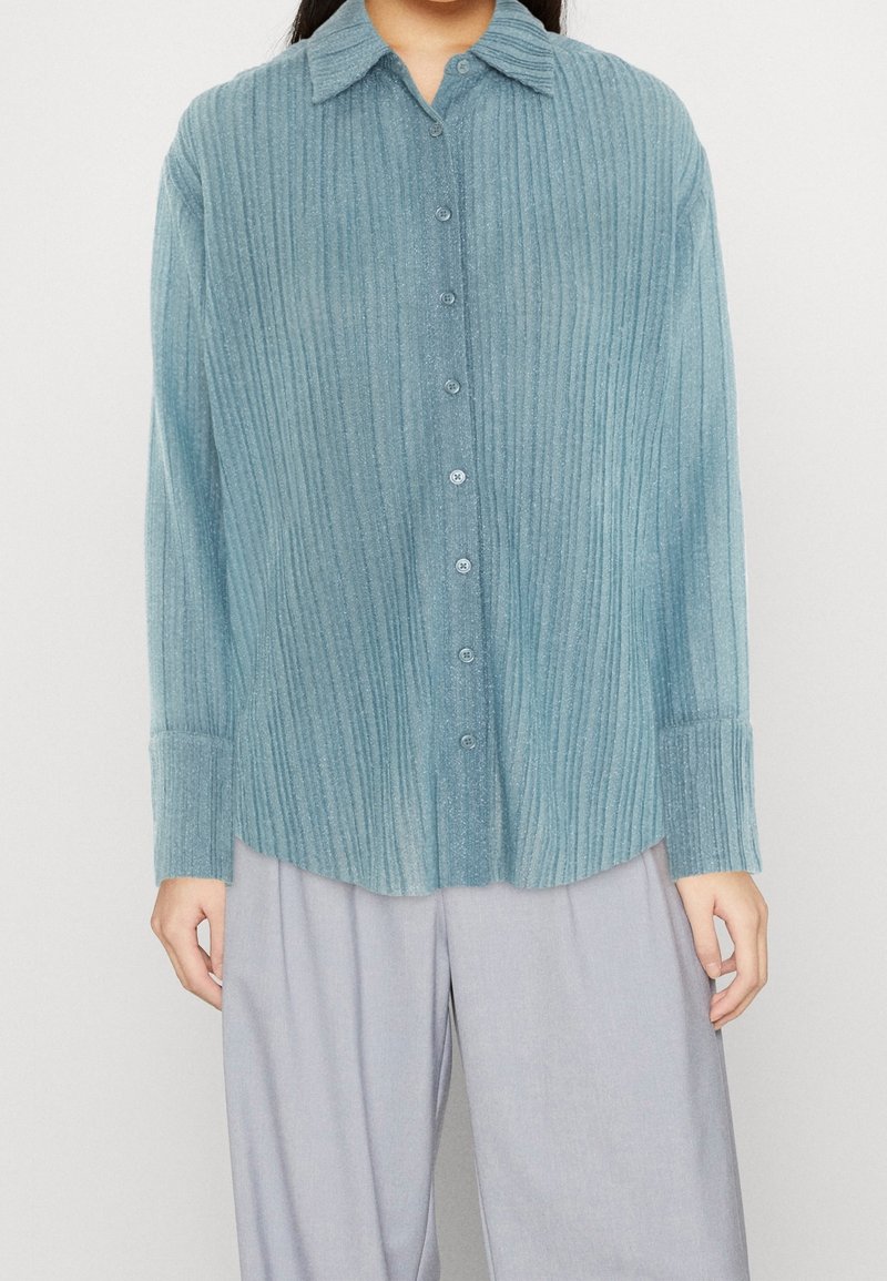 Niebieska koszula zapinana na guziki, plisowana, wykonana z miękkiego, teksturowanego materiału, z kołnierzykiem i długimi rękawami. Posiada pasujące guziki oraz zaokrąglony dół.