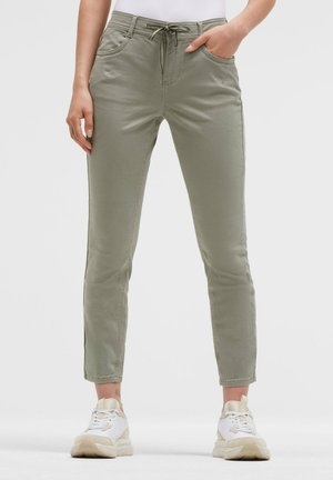 Person, die hellgrüne Slim-Fit-Hosen mit Kordelzug an der Taille und weiße Sneaker trägt, steht vor einem einfarbigen Hintergrund.