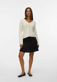 Pull crème à col en V avec fermeture à boutons, associé à une jupe noire évasée. Des ballerines noires avec des accents métalliques complètent la tenue.
