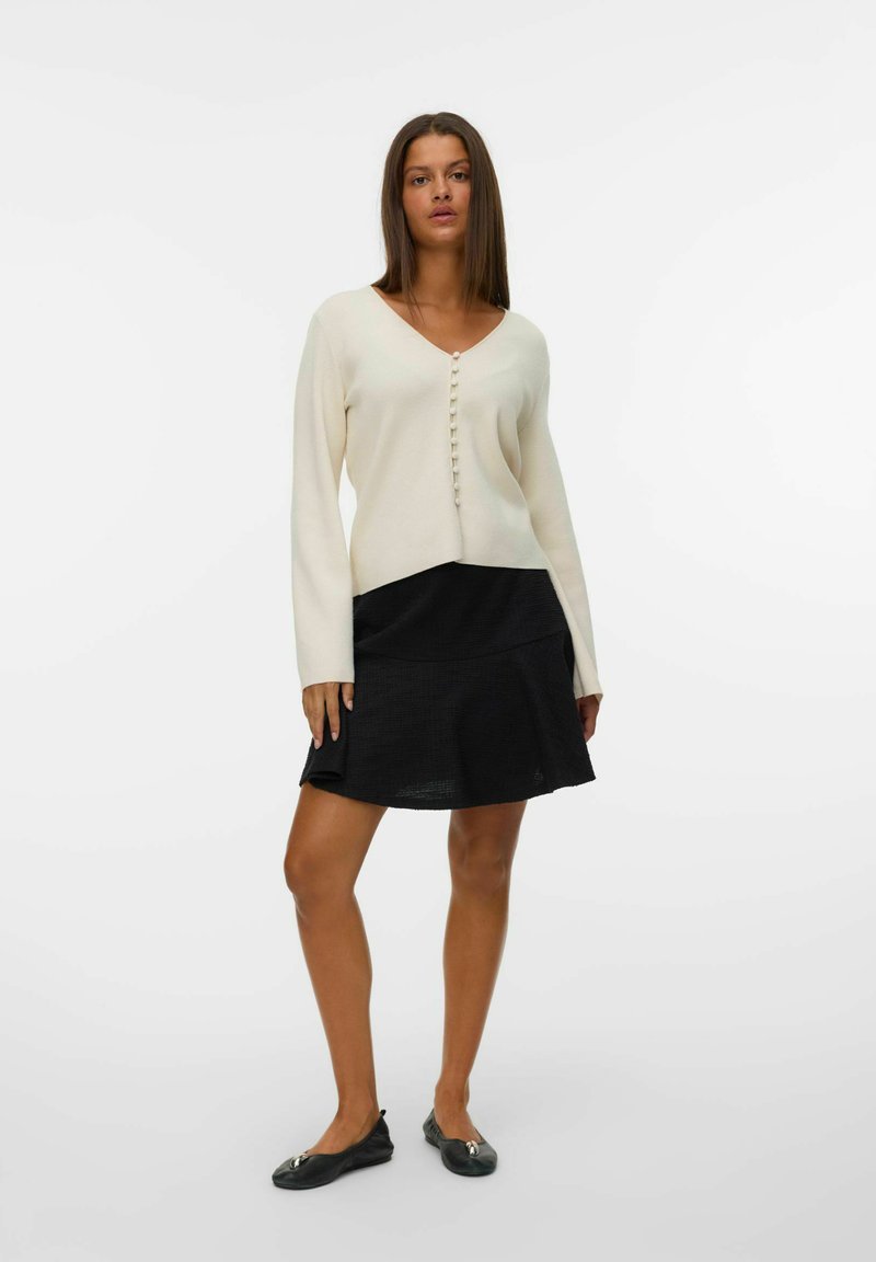 Pull crème à col en V avec fermeture à boutons, associé à une jupe noire évasée. Des ballerines noires avec des accents métalliques complètent la tenue.