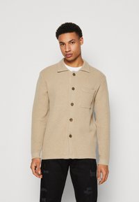 Cardigan beige texturé avec un col rabattu, devant à boutons, poche poitrine unique et boutons marron. Porté avec un pantalon noir déchiré.