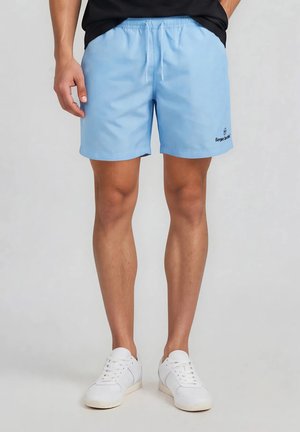 Mand iført lyseblå afslappede shorts med elastisk talje og hvide sneakers, stående mod ensfarvet baggrund.