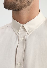 Beige overhemdd met een Mandarin-kraag, voorzien van witte knopen en een zachte, gestructureerde stof. Close-up van de halslijn.