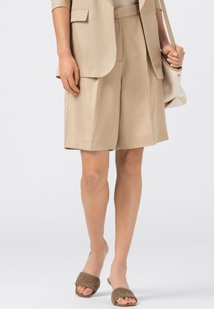 Femme portant un short beige sur mesure, un blazer assorti, des sandales tissées à bout ouvert, et portant un sac à main beige à l'épaule.