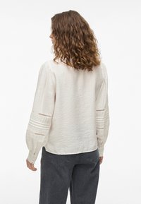 Bluză de culoare crem deschis, dintr-un material texturat, cu mâneci lungi tip balon, care au pliseuri orizontale și accente de dantelă la tivul mânecilor, asortată cu blugi negri.