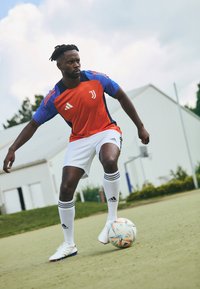 Camisola desportiva vermelha e azul com mangas curtas, calções brancos e meias brancas com riscas pretas; jogador a driblar uma bola de futebol multicolorida.
