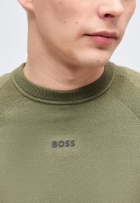Gros plan sur une personne portant un pull en maille vert olive avec le mot "BOSS" sur la poitrine, montrant le bas du visage et le cou.