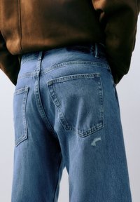 Blauwe spijkerbroek met hoge taille en een ontspannen pasvorm, voorzien van twee achterzakken en lichte slijtage, afgebeeld tegen een neutrale achtergrond.