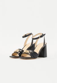 Zwarte leren sandalen met hak, open teen, enkelbandje en gouden hardware detail aan de voorkant, voorzien van een chunky blokhak.