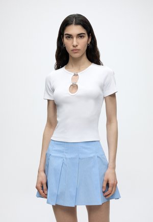 Γυναίκα με μακριά σκουρόχρωμα μαλλιά φοράει λευκό κοντομάνικο τοπ με δύο διακοσμητικά ανοίγματα μπροστά και ανοιχτό μπλε πλισέ φούστα με ρίγες.