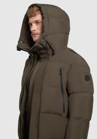 Pružná bunda v tmavě olivově zelené barvě, vybavená velkou kapucí, zapínacími kapsami a logem na levém rukávu. Hladký, matný povrch.