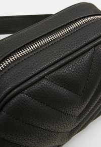 Musta teksturoitu nahkalaukku, jossa hopeanvärinen metallivetoketju ja chevron-pistetyö koristeena pinnassa.