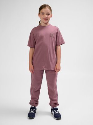 Ung pige står smilende, iført en lilla oversized t-shirt og matchende bukser med mørkeblå og hvide sneakers på en ensfarvet baggrund.
