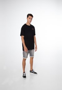 Zwarte katoenen t-shirt met korte mouwen, met een klein rood geborduurd logo. Gecombineerd met lichtgrijze opgerolde shorts en zwarte sneakers.