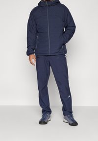 Blå isolerad jacka med huva, kombinerad med matchande marinblå byxor, har zipfickor och texturerad tyg. Skorna är grå med blå accenter.