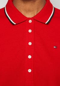 Röd polotröja med en enhetlig struktur, med en krage i svart och vitt randigt mönster, fem vita knappar och en broderad logotyp i hörnet.