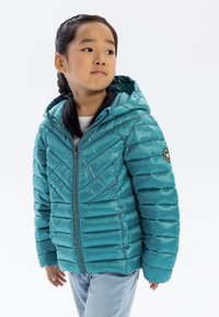 Teal vadderad jacka med chevronmönster, justerbar huva, framdragkedja och logotyp-patch på vänster ärm. Bärs över en vit topp.