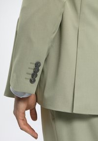 Gros plan sur la main d'une personne près du poignet d'une veste de costume vert clair avec quatre boutons foncés et un pantalon assorti.