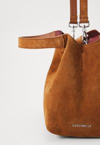 Brown suede bucket bag with two top handles, silver hardware, and a visible brand logo. Soft texture and rounded shape.