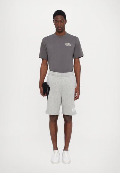Grå t-shirt med logo på brystet, parret med grå shorts med elastisk talje. Modellen har hvide sneakers på og holder en sort genstand.