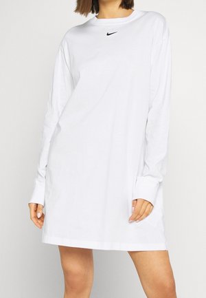 Femme portant une robe blanche à manches longues de Nike avec un petit logo noir sur la poitrine, debout devant un fond clair uni.