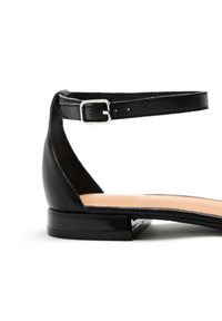 Zapato plano de piel negra con una delgada correa en el tobillo y hebilla plateada, con punta en forma de almendra y tacón bajo. Textura suave.
