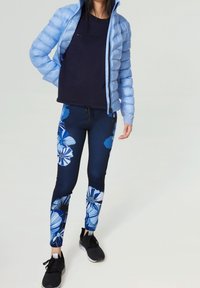 Ljusblå pufferjacka över en marinkolsvit t-shirt, kombinerad med blommiga leggings med blåa och vita mönster, samt svarta träningsskor.