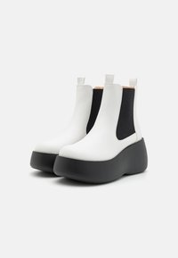 Botas de tobillo de cuero blanco grueso con paneles elásticos negros a los lados, suela plana tipo plataforma y tiradores para facilitar su uso. Textura suave y diseño moderno.