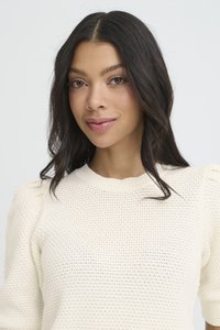 Junge Frau mit langen dunklen Haaren, die einen weißen, strukturierten Strickpullover trägt und sanft lächelt, vor einem schlichten hellen Hintergrund.