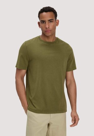 Man die een olijfgroene T-shirt met korte mouwen en beige broek draagt, staand met één hand in de zak tegen een effen achtergrond.