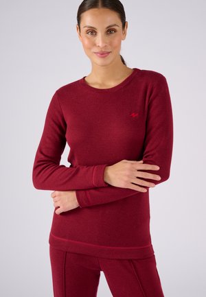 Donna con capelli scuri raccolti in una coda bassa, che indossa una maglia aderente a maniche lunghe color borgogna e pantaloni abbinati, in piedi con le braccia incrociate.