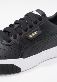 Czarne skórzane sneakersy z perforowaną teksturą, kontrastującą białą podeszwą, czarnymi sznurówkami i złotym detalem logo z boku.