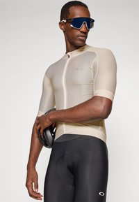 Athlet trägt ein beige Radtrikot, schwarze Radhosen und reflektierende Sonnenbrille und hält einen schwarzen Fahrradhelm vor einem einfarbigen Hintergrund.