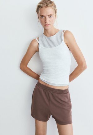 Fiatal nő áll egy fehér fal előtt, szürke réteges ujjatlan felsőt és barna elasztikus derékú rövidnadrágot visel, kezei a háta mögött.