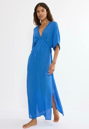 Frau steht barfuß und trägt ein langes blaues Kleid mit V-Ausschnitt, ellbogenlangen Ärmeln und einem Seitenschlitz vor weißem Hintergrund.