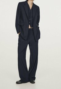 Marinblått randigt kostym med en skräddarsydd kavaj med spårade slag, en välsittande väst och vida byxor. Slät tyg, polerade skor.