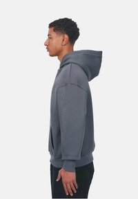 Grauer Hoodie mit einer Fronttasche, Kordelzugkapuze und gerippten Bündchen. Das Material wirkt weich und hat eine entspannte, oversized Passform.