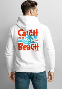 Weißer Kapuzenpullover mit grafischem Design: roter Text "Fang mich am Strand", blaue Wellen, eine Palme und ein kreisförmiges Akzent.
