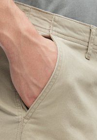 Pantalones cortos de tela beige con un bolsillo lateral, con costuras planas y una textura suave. Una mano se introduce en el bolsillo, mostrando su funcionalidad.