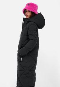 Cappotto piumino nero con un design lungo e trapuntato, cappuccio e tasche con zip. Indossato con un cappello a secchiello rosa e peloso con un accento del logo.