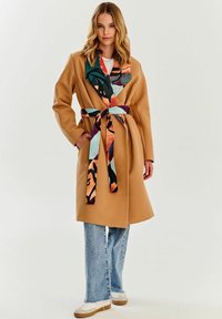 Beżowy płaszcz z materiału z wielokolorowym wzorzystym kołnierzem i paskiem. Noszony na białej bluzce, zestawiony z jasnymi dżinsami i białymi trampkami.