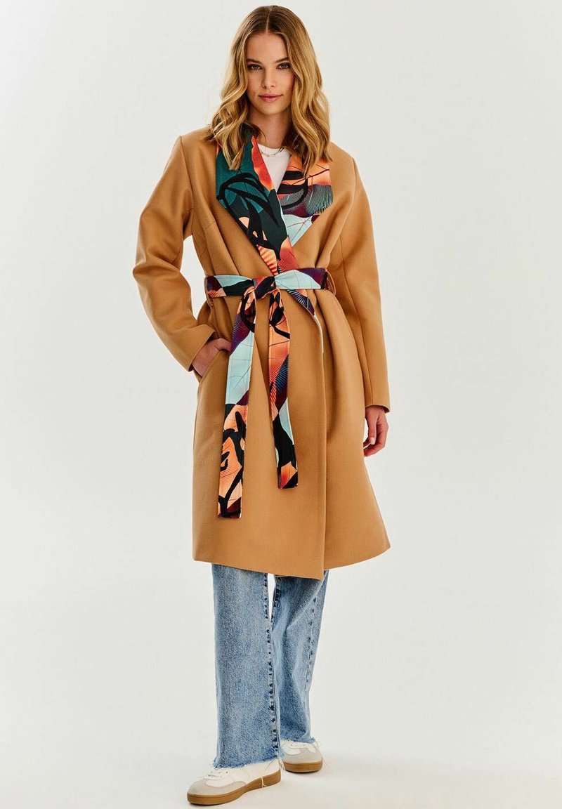 Beżowy płaszcz z materiału z wielokolorowym wzorzystym kołnierzem i paskiem. Noszony na białej bluzce, zestawiony z jasnymi dżinsami i białymi trampkami.