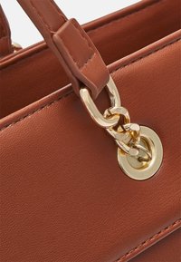 Sac à main en cuir marron présentant une texture lisse, des accessoires en or et un attachement en chaînes près d'un œillet en métal circulaire.