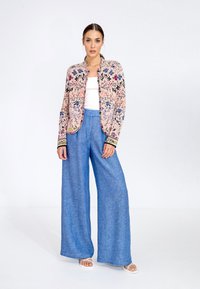 Floral-patterned, long-sleeve jacket in pink and blue, paired with loose light blue linen trousers. Model wears a white top and sandals.