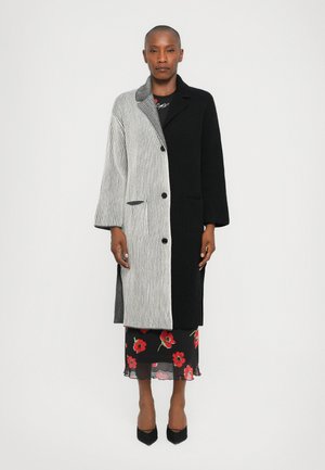 Manteau en tricot avec un design rayé noir et gris, doté d'un col, de boutons et de poches latérales. Porté par-dessus une robe nuisette à fleurs et avec des talons.