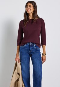 Lächelnde Frau in einem dunkelvioletten Langarmoberteil und blauer Jeans, die einen beigen Trenchcoat in der linken Hand hält.